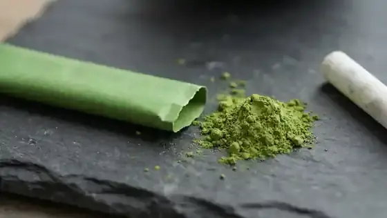 A slim green paper stick packet of matcha torn open at the top beside a small mound of bright green powder on a dark slate surface